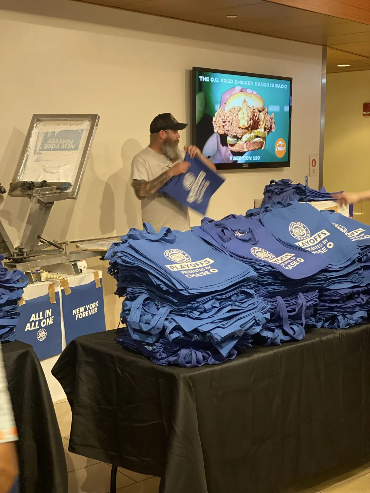 Stacks of printed Knicks Playoffs totes on the table while the crew prints at an arena concourse station
