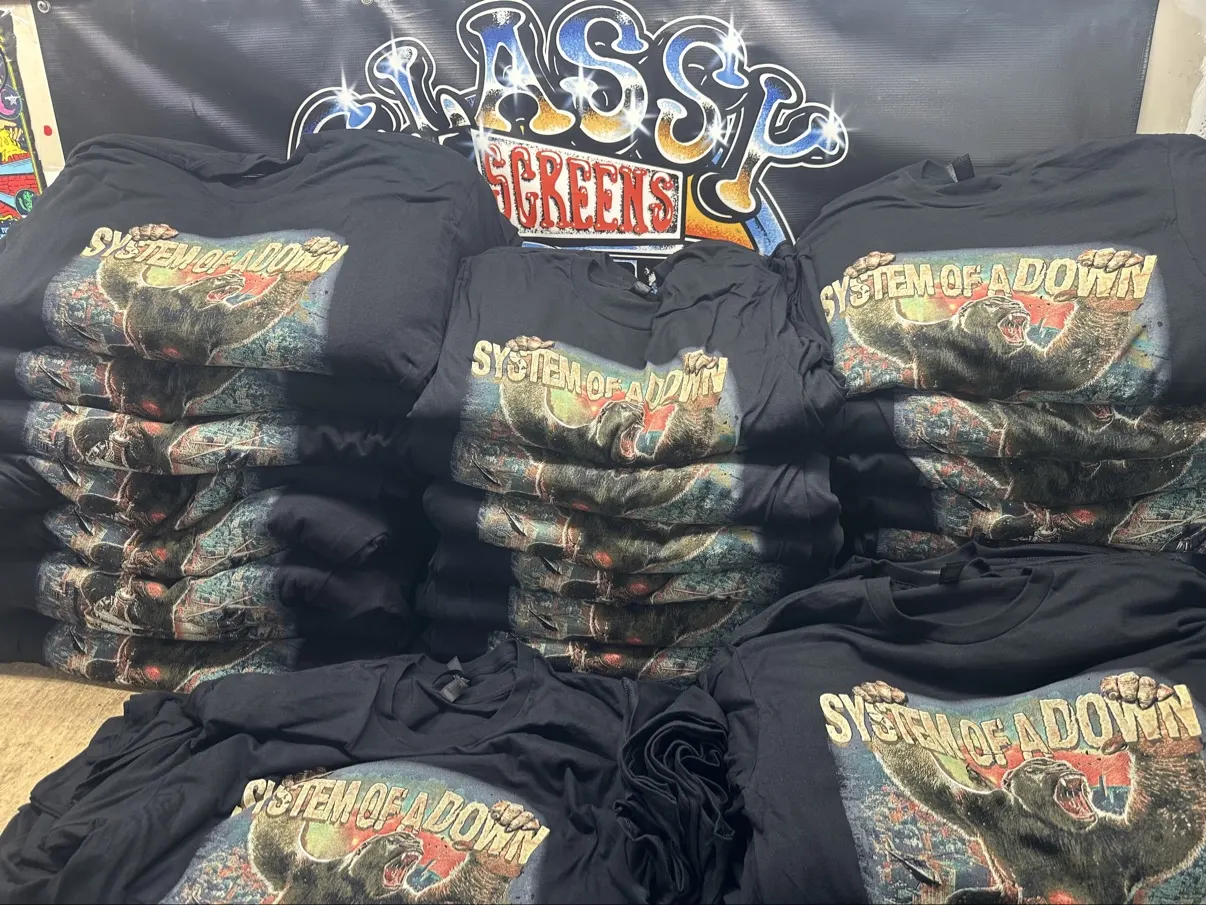 Stacks of System of a Down MetLife Stadium tees in front of the Classy Screens banner